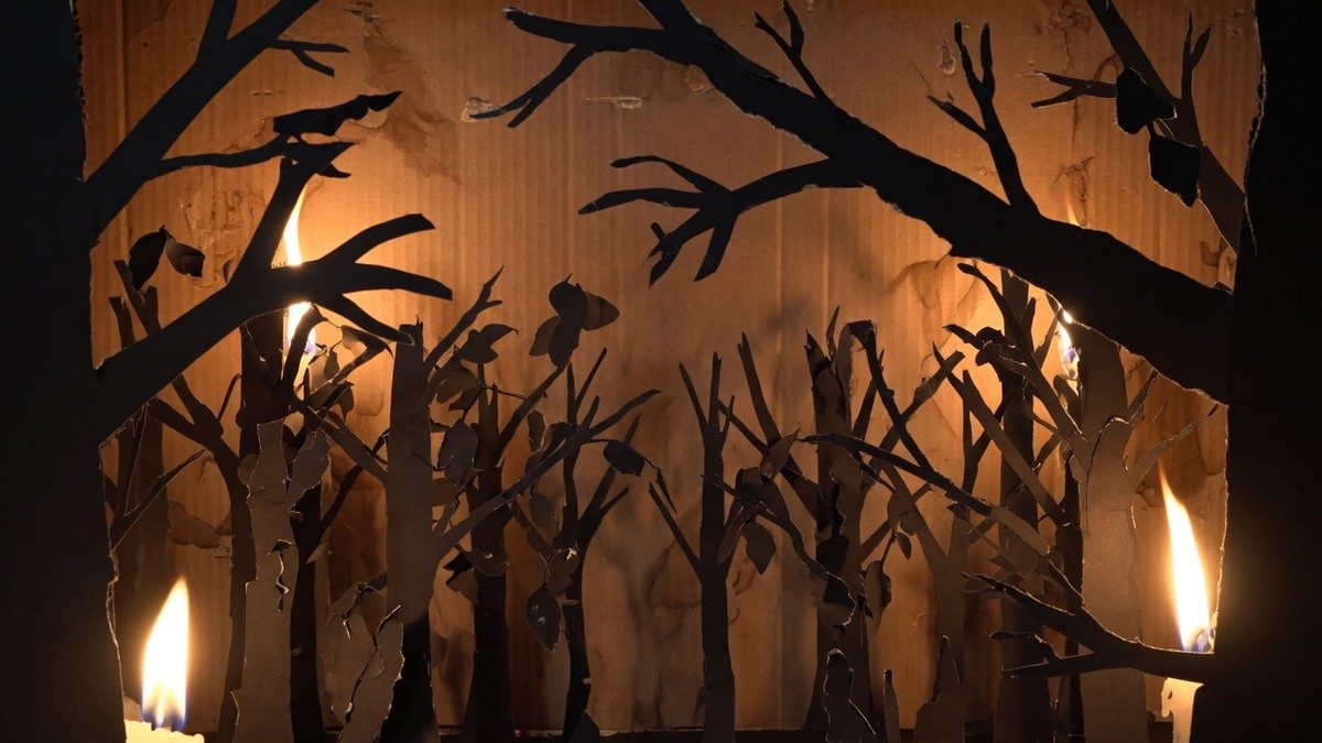 Décor de forêt en papier découpé et carton, éclairé par des flammes projetant des ombres inquiétantes, dans une ambiance de conte de fées sombre