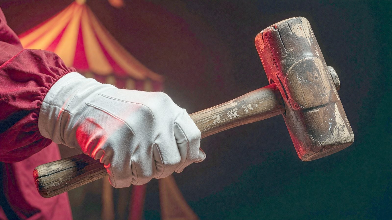 Gros plan sur une main gantée de blanc serrant un vieux maillet en bois, éclairée par une lumière rouge de fête foraine.