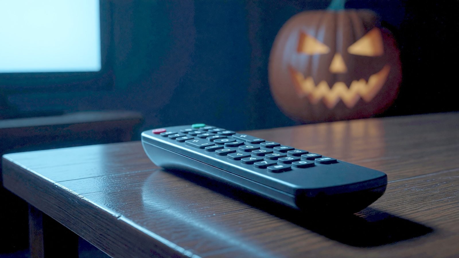 Une télécommande de télévision posée sur une table basse, éclairée par la lueur bleutée d'un écran dans l'obscurité, avec une citrouille d'Halloween en arrière-plan flou