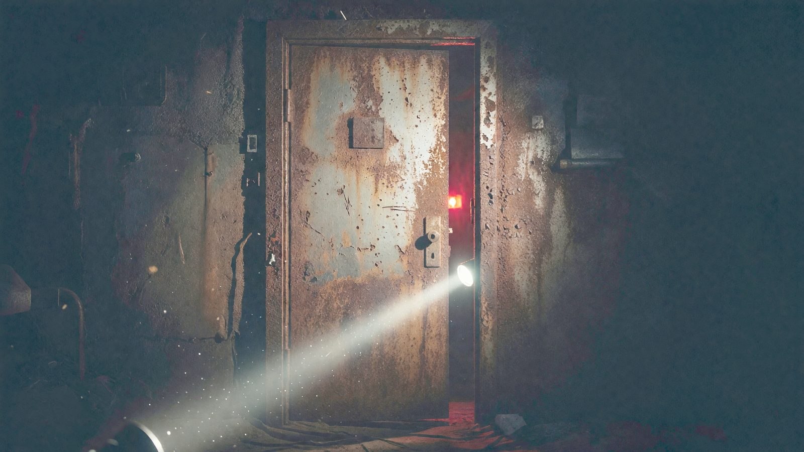 Porte métallique de sous-sol entrouverte, traversée par un faisceau de lampe torche dans la pénombre