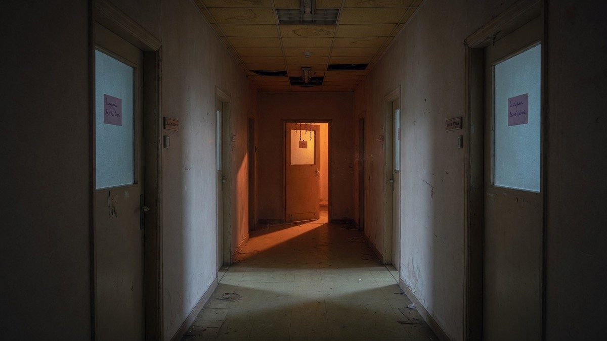 Couloir sombre et silencieux d'un bâtiment évoquant une secte, avec une porte entrouverte au fond laissant filtrer une lumière inquiétante