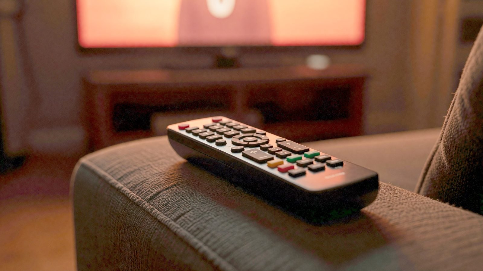 Gros plan sur une télécommande posée sur un canapé, éclairée par la lueur rouge orangé d'un écran de télévision hors champ