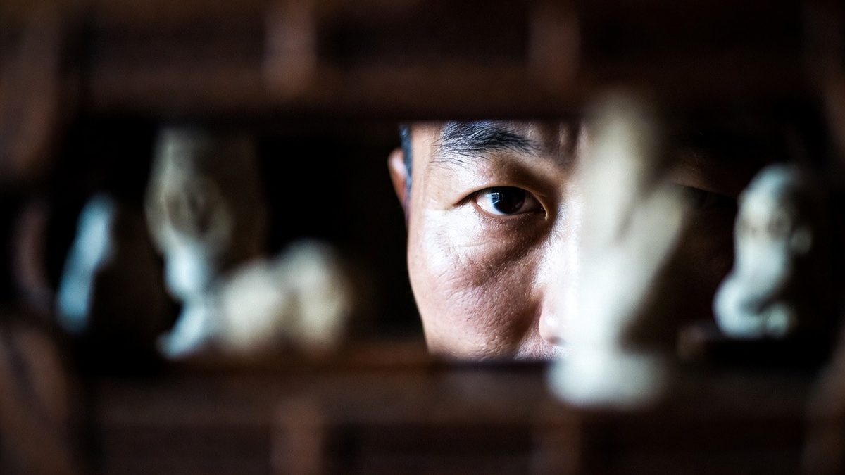 Portrait d’un homme asiatique regardant à travers une ouverture sculptée en bois, expression intense et mélancolique – illustration de The Ugly de Yeon Sang-ho