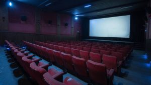 Salle de cinéma vide baignée dans une lumière bleue froide, fauteuils rouges alignés face à un grand écran blanc lumineux