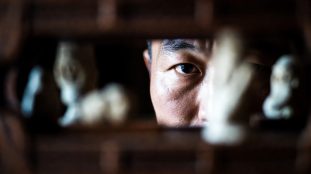 Portrait d’un homme asiatique regardant à travers une ouverture sculptée en bois, expression intense et mélancolique – illustration de The Ugly de Yeon Sang-ho