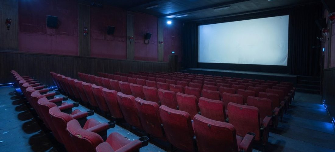 Salle de cinéma vide baignée dans une lumière bleue froide, fauteuils rouges alignés face à un grand écran blanc lumineux